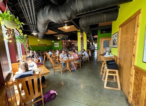 A vibrant restaurant interior with wooden tables, patrons dining, and green walls, featuring plants and industrial decor.