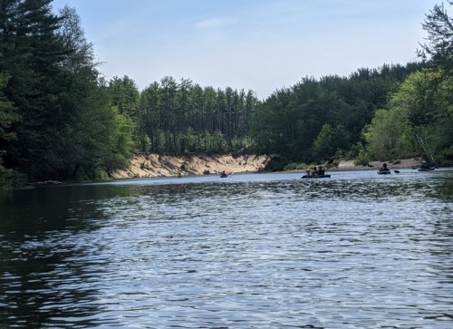 A serene river scene with kayakers paddling, surrounded by lush green trees and a sandy bank in the background.