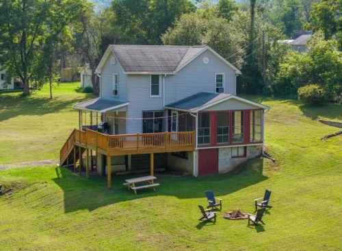 A two-story house with a wooden deck and screened porch, surrounded by green grass and trees. Fire pit and chairs in front.