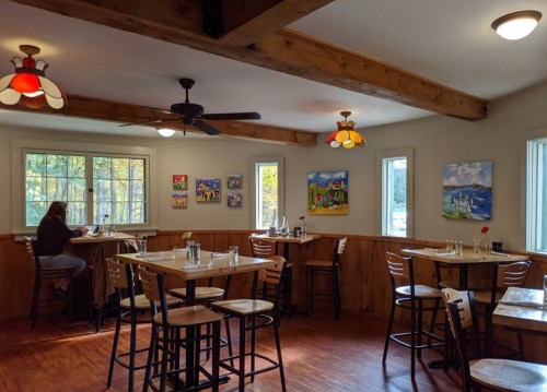Cozy restaurant interior with wooden beams, tables, and colorful artwork on the walls. A person sits at a table.