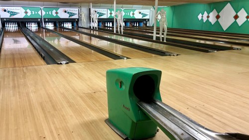 A bowling alley with empty lanes, a green ball return, and white mannequins positioned at the end of the lanes.