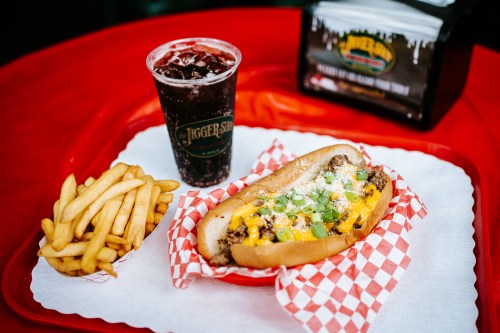 A hot dog topped with cheese and green onions, served with fries and a soda on a red table.