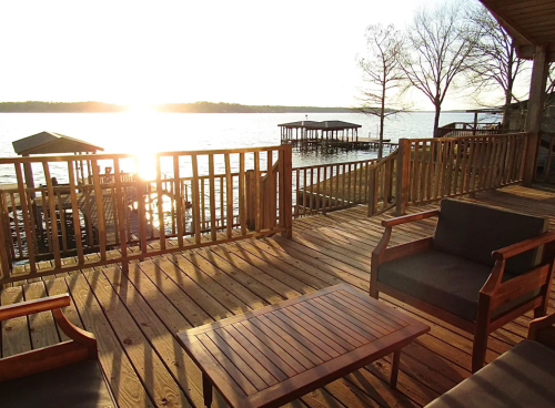 A wooden deck overlooking a lake at sunset, with chairs and a table, and docks in the background.