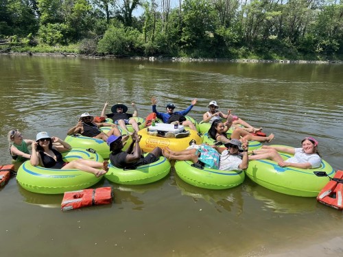 A group of people in green inner tubes floating on a river, enjoying a sunny day with smiles and hats.