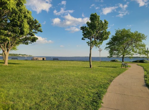 A serene park scene with green grass, trees, a walkway, and a view of the water under a blue sky with clouds.