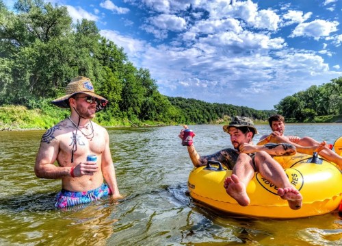 Three friends enjoy a sunny day on the river, one in a yellow inner tube, holding drinks, surrounded by greenery.