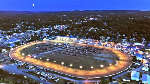 Aerial view of a racetrack at dusk, surrounded by a fair with lights, cars, and a moonlit sky.