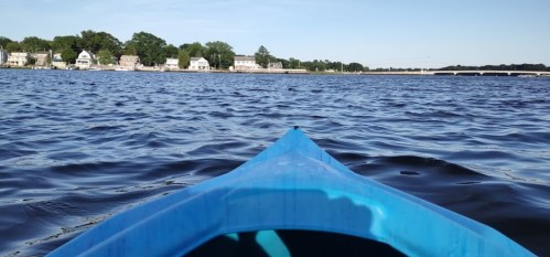 View from a kayak on calm water, with a shoreline of houses and trees in the background under a clear blue sky.