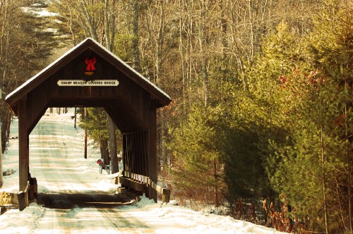 A snow-covered road leads to a wooden covered bridge surrounded by trees in winter.