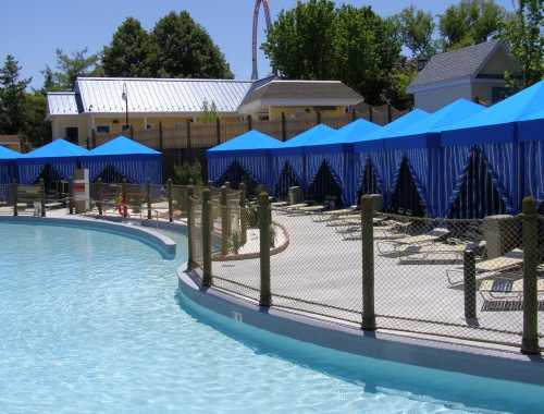 A sunny pool area with blue cabanas, lounge chairs, and a curved water edge, surrounded by a fence and greenery.