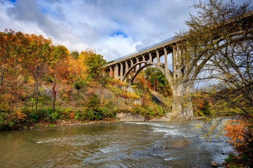 A scenic view of a bridge arching over a river, surrounded by vibrant autumn foliage and a cloudy sky.