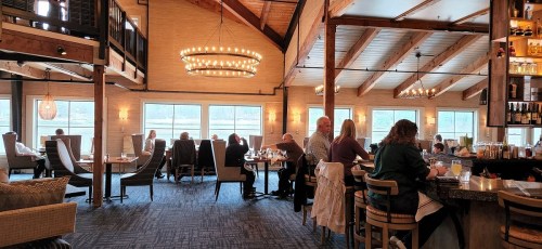 A cozy restaurant interior with diners at tables and a bar, featuring large windows and wooden beams.