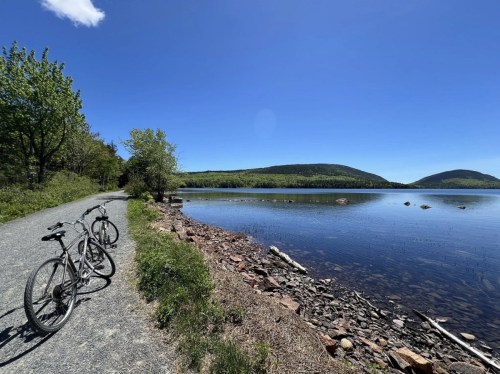 Two bicycles parked on a gravel path beside a calm lake, surrounded by lush greenery and distant hills under a clear blue sky.