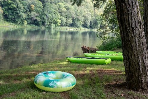 A green inflatable tube and kayaks by a calm river, surrounded by trees and lush greenery.