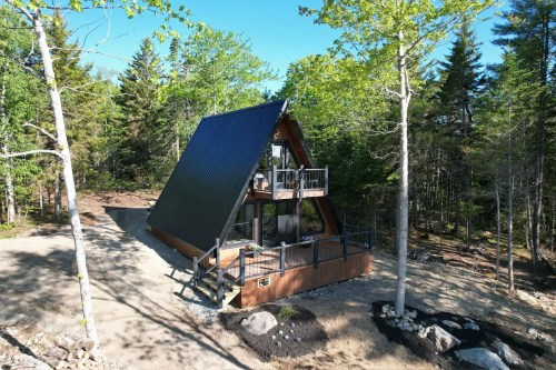 A modern A-frame cabin surrounded by trees, featuring a deck and a gravel pathway in a serene outdoor setting.