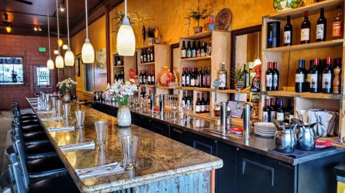 A stylish bar with a long countertop, shelves of wine and spirits, and floral arrangements in a warm, inviting setting.