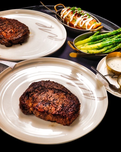 Two plates of steak with sides of asparagus and a loaded baked potato, elegantly presented on a dark table.