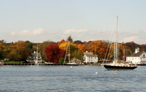 A sailboat on calm water with colorful autumn trees and charming houses in the background.