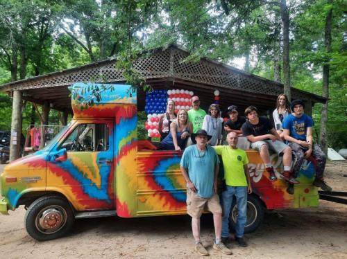 A colorful van with a tie-dye design parked in front of a wooden structure, surrounded by a group of people posing together.