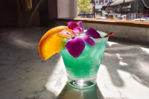 A vibrant blue cocktail garnished with an orange slice and a purple flower, sitting on a marble surface.