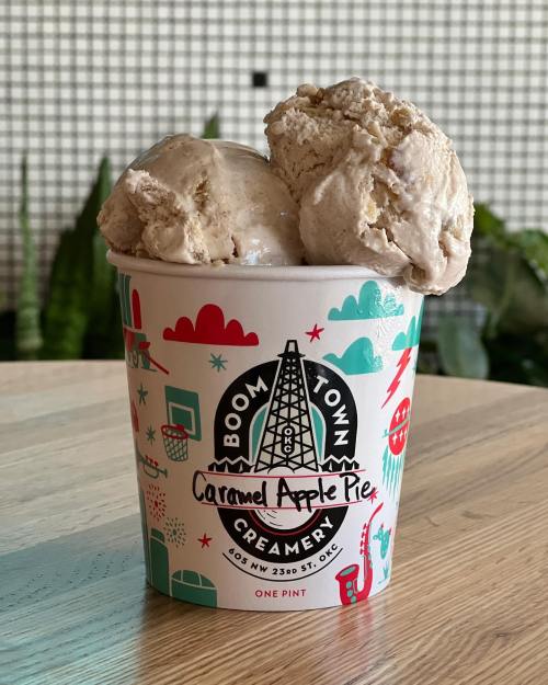 A pint of caramel apple pie ice cream with two scoops, in a colorful container, on a wooden table.