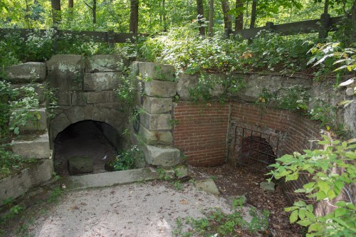Two stone archways partially covered by greenery, leading into a wooded area. One archway has a metal grate.