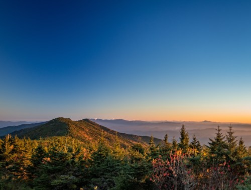 A scenic mountain view at sunset, with lush green trees and a clear blue sky fading into warm hues.