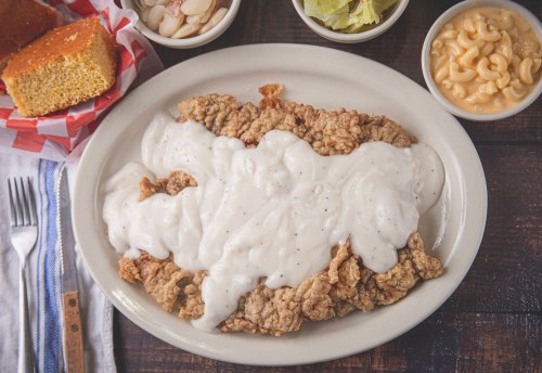 A plate of fried chicken smothered in white gravy, served with cornbread, macaroni, and sides of salad and pickles.