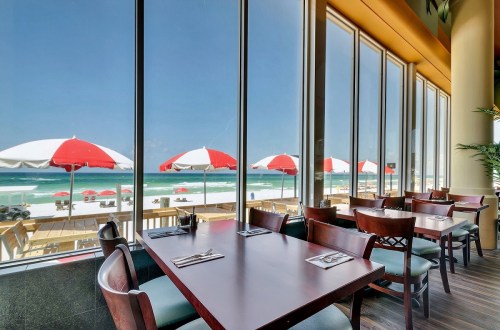 A beachside restaurant with large windows, showcasing red and white umbrellas on the sandy shore and turquoise water.