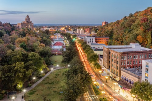 Aerial view of a city at dusk, featuring a tree-lined street, buildings, and lights illuminating the scene.