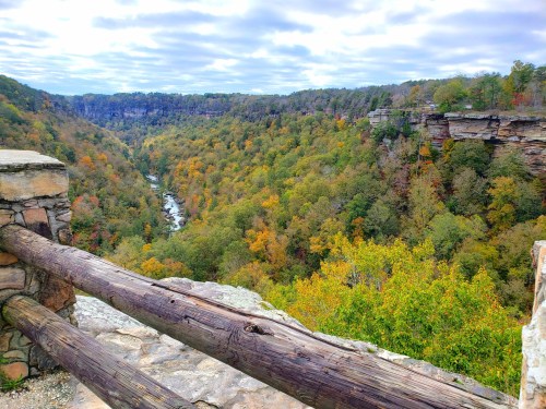 A scenic view of a colorful forested valley with a river, framed by rocky cliffs under a cloudy sky.