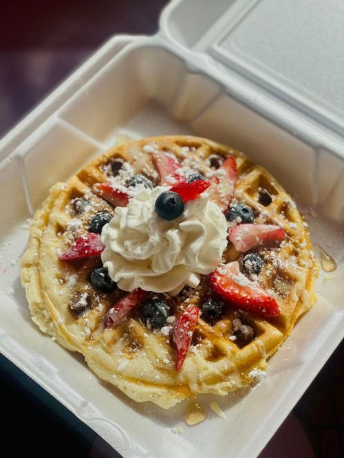 A delicious waffle topped with strawberries, blueberries, whipped cream, and a drizzle of syrup in a takeout container.