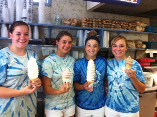 Four young women in tie-dye shirts hold large ice cream cones in a shop, smiling at the camera.