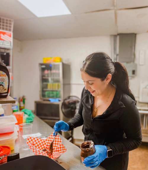 A woman in a black shirt and blue gloves smiles while preparing food in a kitchen, holding a jar of chocolate spread.