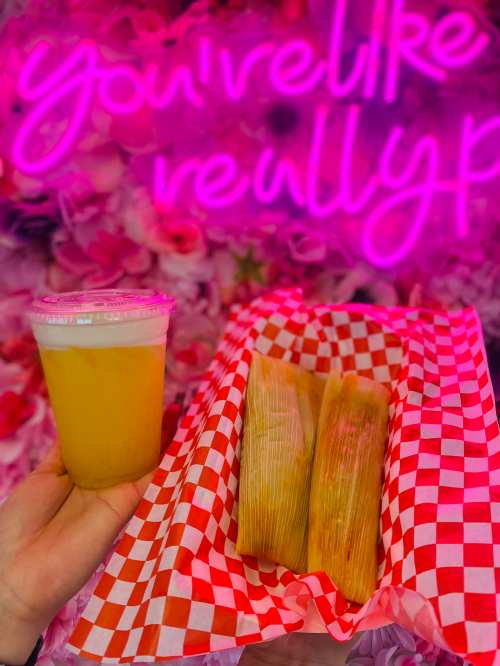 A hand holding a drink and a tray of tamales in front of a pink floral backdrop with neon text.