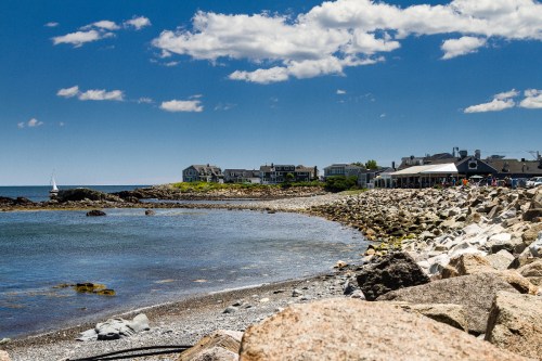 A rocky shoreline with a calm sea, dotted with houses and a sailboat under a bright blue sky with fluffy clouds.