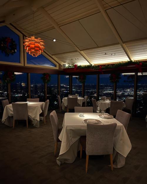 Elegant restaurant interior with tables set for dining, large windows showcasing a city skyline at night, festive decorations.
