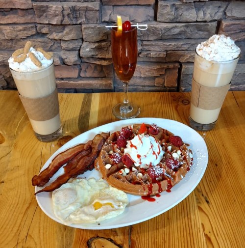 A plate of waffles topped with whipped cream, strawberries, and syrup, accompanied by bacon, eggs, and two drinks.