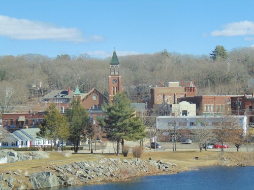 A scenic view of a small town with a clock tower, trees, and a lake under a clear blue sky.