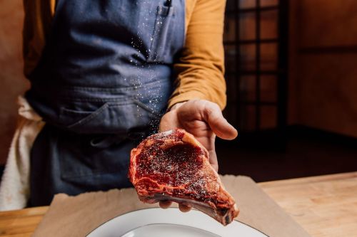 A person in an apron sprinkles salt over a raw steak held above a plate on a wooden table.