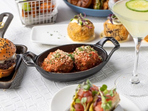 A close-up of meatballs in marinara sauce, garnished with herbs, alongside a cocktail and various appetizers.