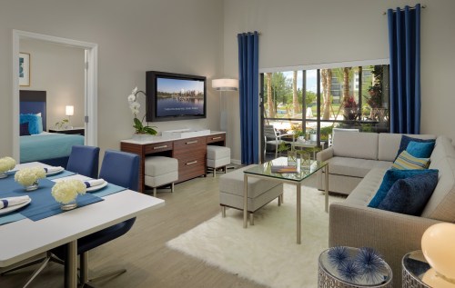Modern living room with a sofa, glass coffee table, and dining area, featuring blue accents and large windows.