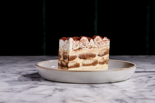 A slice of layered tiramisu cake on a round plate, set against a dark background.