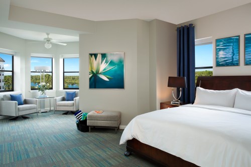 Bright hotel room featuring a king bed, seating area, large windows, and modern decor with a floral artwork.