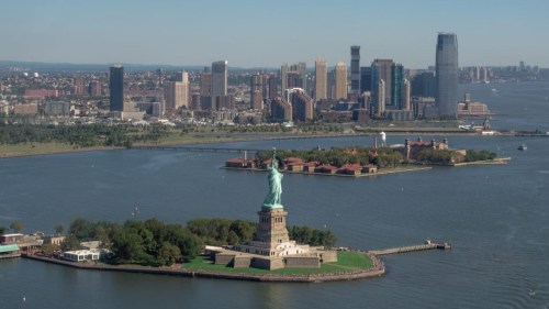 Aerial view of the Statue of Liberty on Liberty Island, with a skyline of buildings in the background.