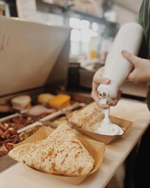 A hand squeezes whipped cream onto a crepe, with two crepes in paper trays on a counter.