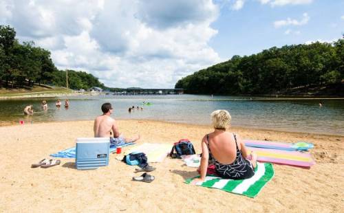 Two people sit on a sandy beach by a lake, with others swimming and enjoying the sunny day in the background.