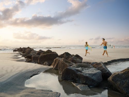 A man and a girl walk along a beach, carrying a surfboard, with rocks and gentle waves in the background.