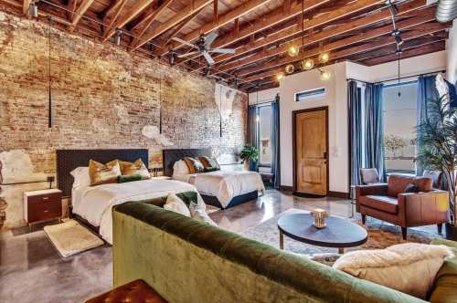 Stylish hotel room with exposed brick walls, two beds, a green sofa, and large windows letting in natural light.
