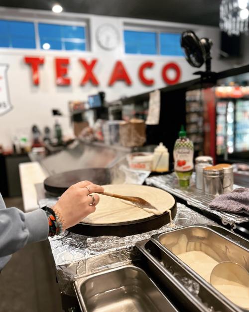 A hand spreads batter on a hot griddle in a diner with a "TEXACO" sign in the background.
